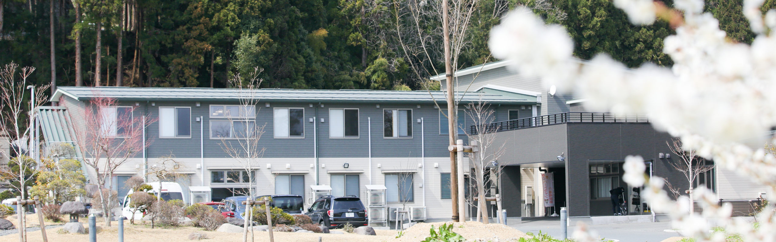 白と濃紺の外壁を持つ介護施設の建物全景。周囲に樹木があり、駐車スペースが見える。